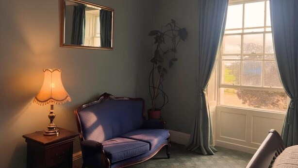 A room with a blue sofa, a bedside table with a lamp, a mirror, and a window with curtains. - South Hill House, Historic Georgian manor with capacity for 52 guests (Delvin)