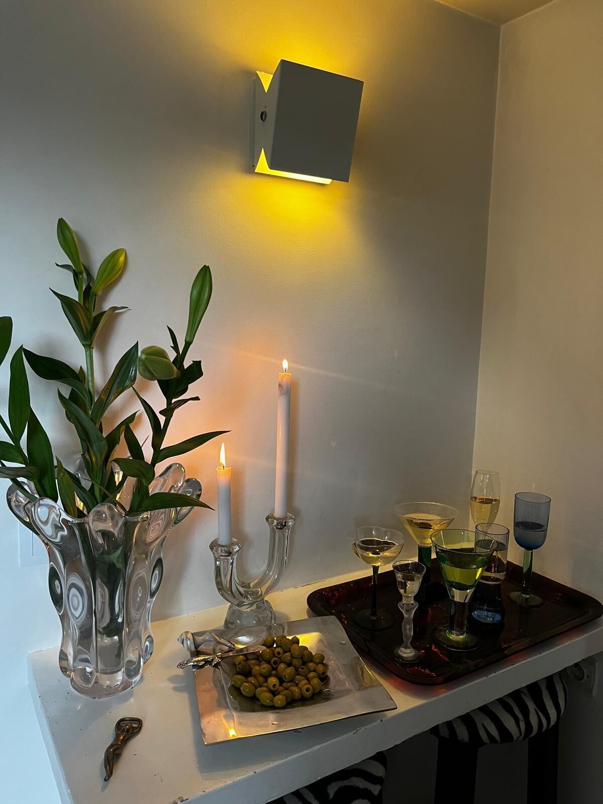 A table with a candle, a glass vase with green leaves, and a tray with olives and glasses.