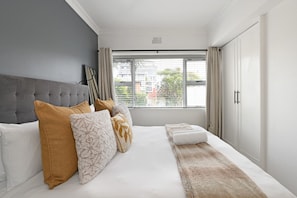 A modern bedroom with a grey headboard, a large bed with white bedding, multiple pillows, and a view of the outside through a window with blinds. - Sea Point 2bed Home Spacious Central Secure (Cape Town)