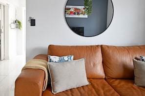 A brown leather sofa with cushions and a throw blanket, a round mirror on the wall, and a small plant on a shelf. - Sea Point 2bed Home Spacious Central Secure (Cape Town)