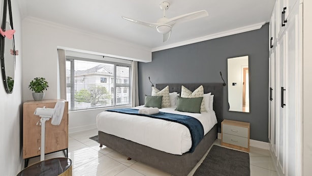 Un dormitorio moderno con una cama, ventilador de techo, ventana, espejo y mesita de noche.