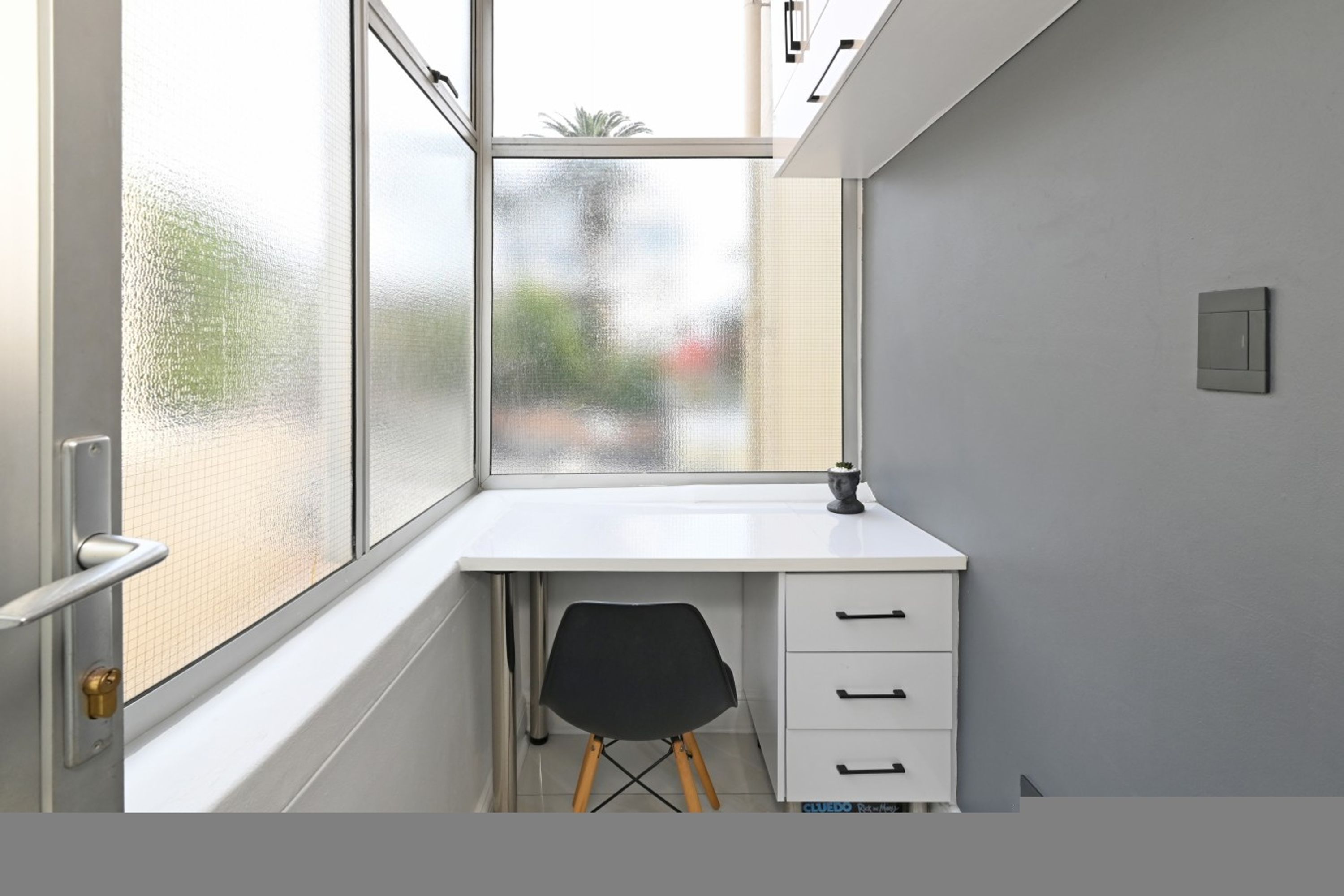 A small, white desk with a chair, a window with a view of trees, and a door with a handle.