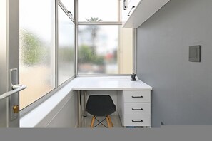 A small, white desk with a chair, a window with a view of trees, and a door with a handle. - Sea Point 2bed Home Spacious Central Secure (Cape Town)