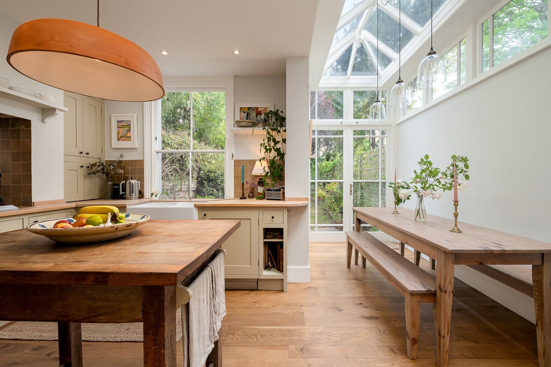 A kitchen with a wooden table, a hanging light fixture, and a view of the garden through large windows.