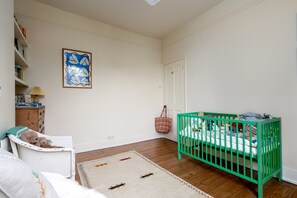 A room with a crib, a chair with a cushion, a small table with a lamp, a wall with a framed picture, and a door. - GuestReady - Stylish Victorian Home with Garden (London)