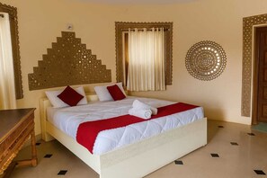 A hotel room with a bed, a wooden side table, a decorative wall piece, and a window with curtains. - Dholavira Tourism Resort  (Dholavira)