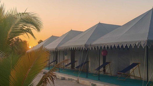 A row of white tents with blue lounge chairs, a swimming pool, and palm trees at sunset.
