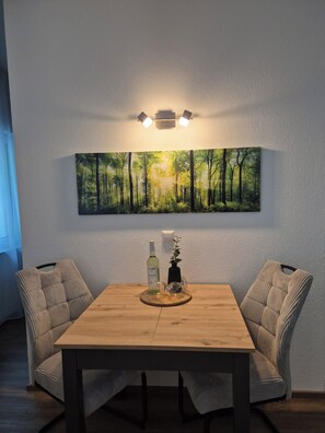 A dining area with a wooden table, two upholstered chairs, a wall-mounted light fixture, and a framed forest landscape painting. - Vrbo Property (Auerbach/Vogtland)