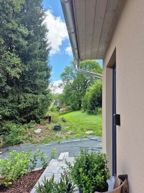 A residential outdoor area with a paved pathway, a small garden, and a house with a door and window.
