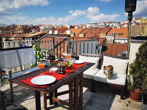 A rooftop terrace with a dining table set for two, a dog sitting on a cushioned bench, and a cityscape view in the background. - Rooftop Terrace Loft Apartment, Home Cinema (Prague)