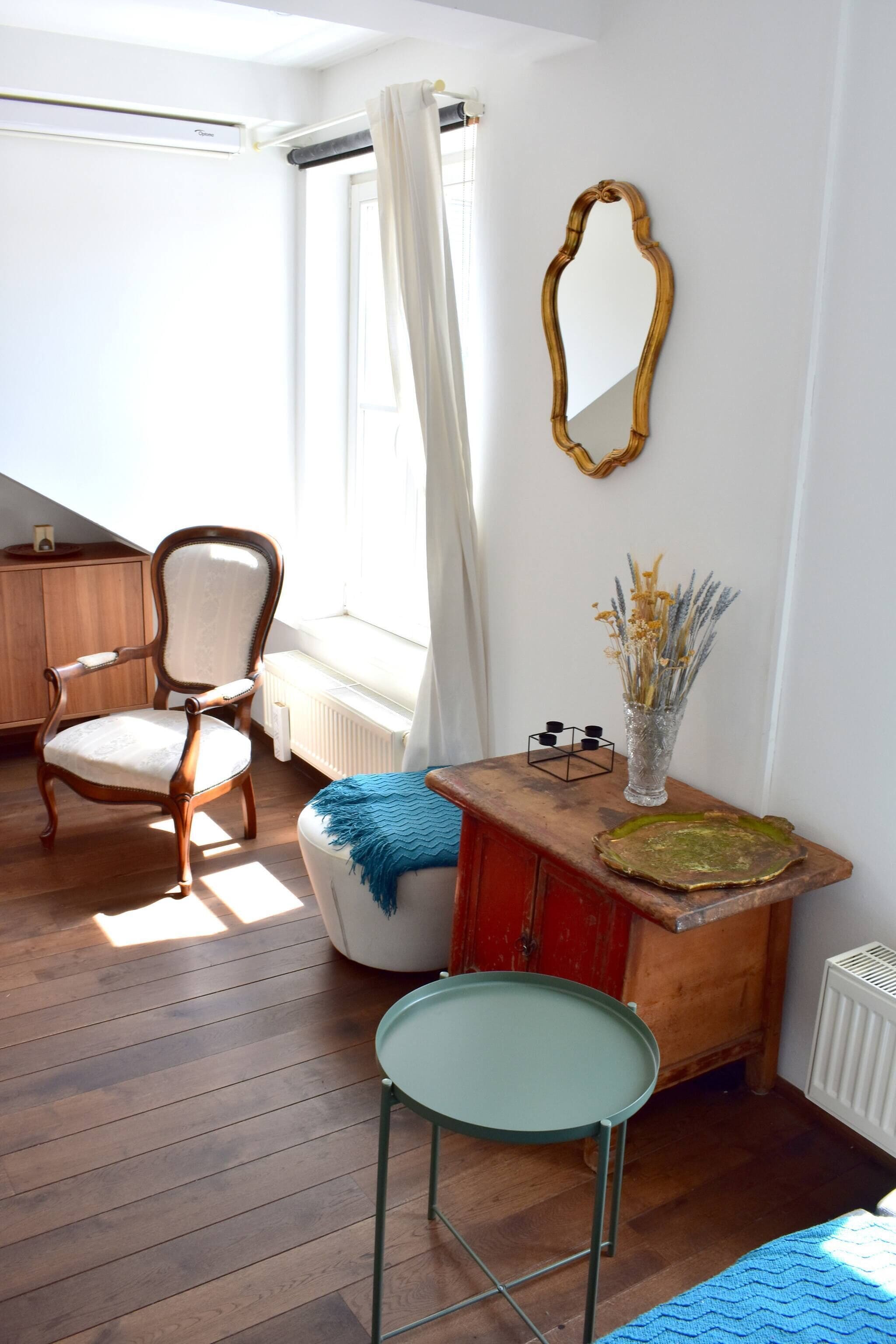 Una habitación con una silla de madera, una mesita con un florero, un espejo en la pared y un radiador.