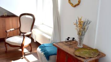 Una habitación con una silla de madera, una mesita con un florero, un espejo en la pared y un radiador.