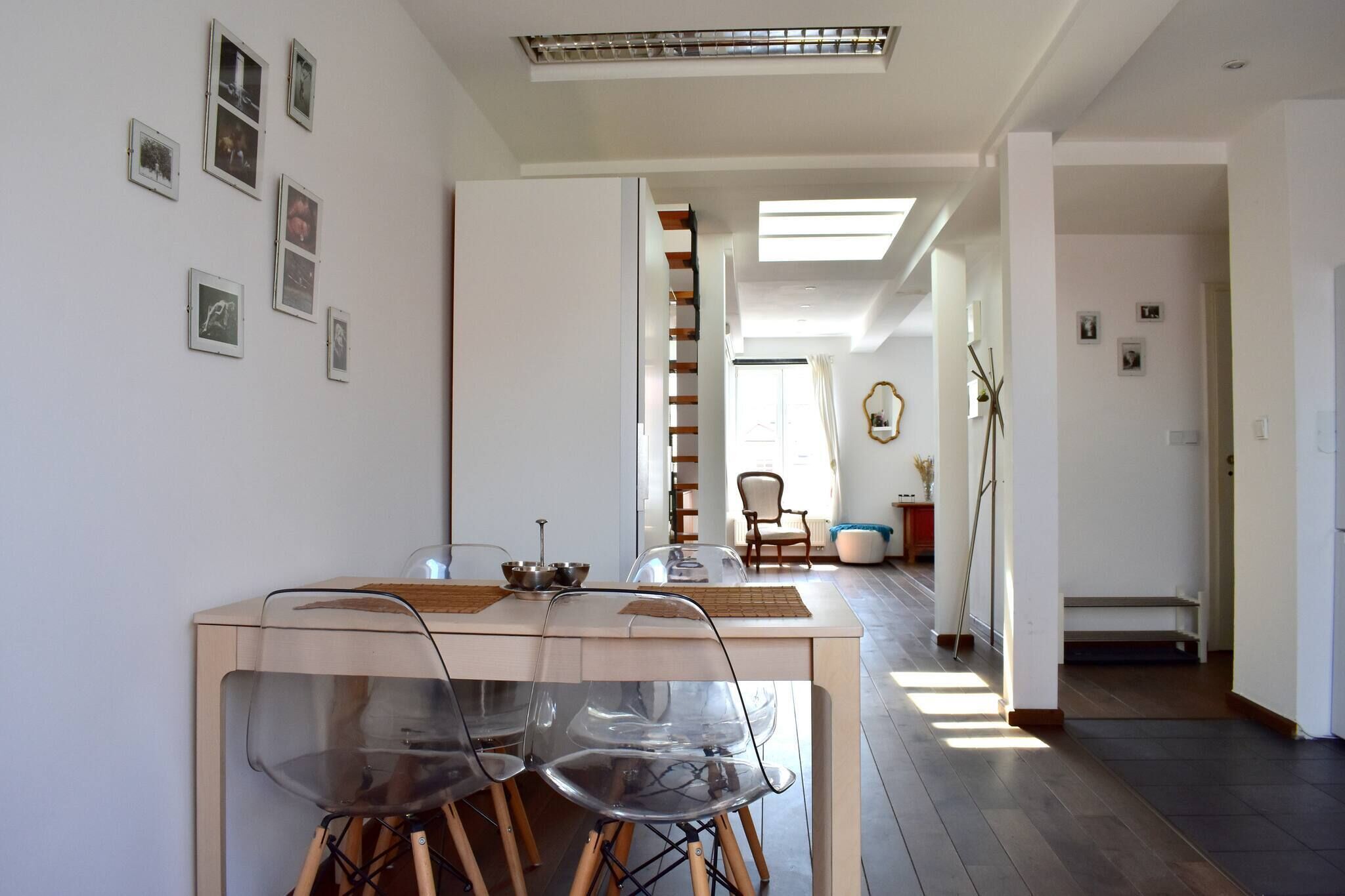 Un interior moderno con zona de comedor que incluye una mesa de madera y sillas traslúcidas, una escalera que sube a otro nivel y un pasillo con un espejo y cuadros en la pared.