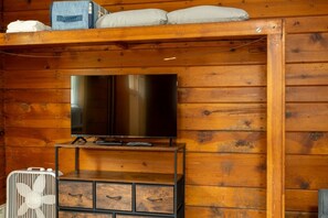 A wooden cabin interior with a television on a stand, a small cabinet below, and a fan on the floor.