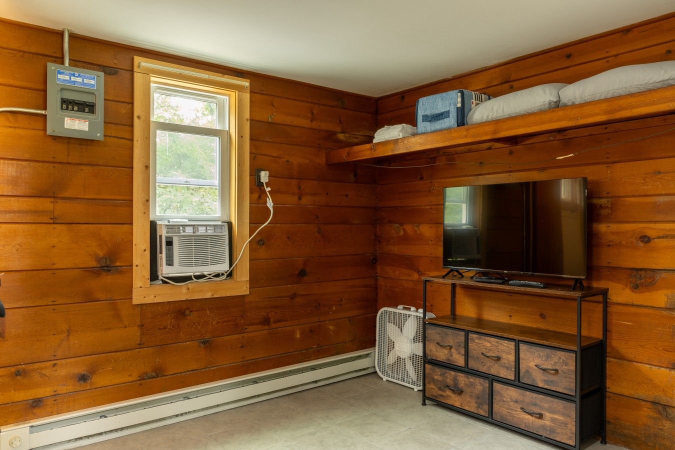 Une pièce aux murs en bois, avec une fenêtre, un appareil de climatisation, un téléviseur posé sur un meuble en bois et un ventilateur.
