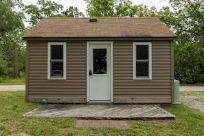 A small, single-story house with a front door, two windows, and a wooden deck.