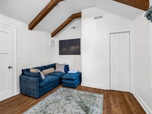 A room with a blue sectional sofa, a patterned rug, wooden flooring, white walls, and exposed wooden beams. - Hendersonville Honeybee (Hendersonville)