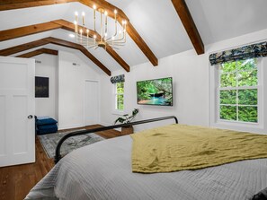 Ein Schlafzimmer mit Bett, einer Chandelüster, Holzbalken, einem Fernseher, einem Fenster mit Blick ins Grüne und einer Tür.
