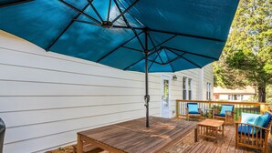 Um deck de madeira com uma mesa e cadeiras, um guarda-sol azul e uma casa branca ao fundo.