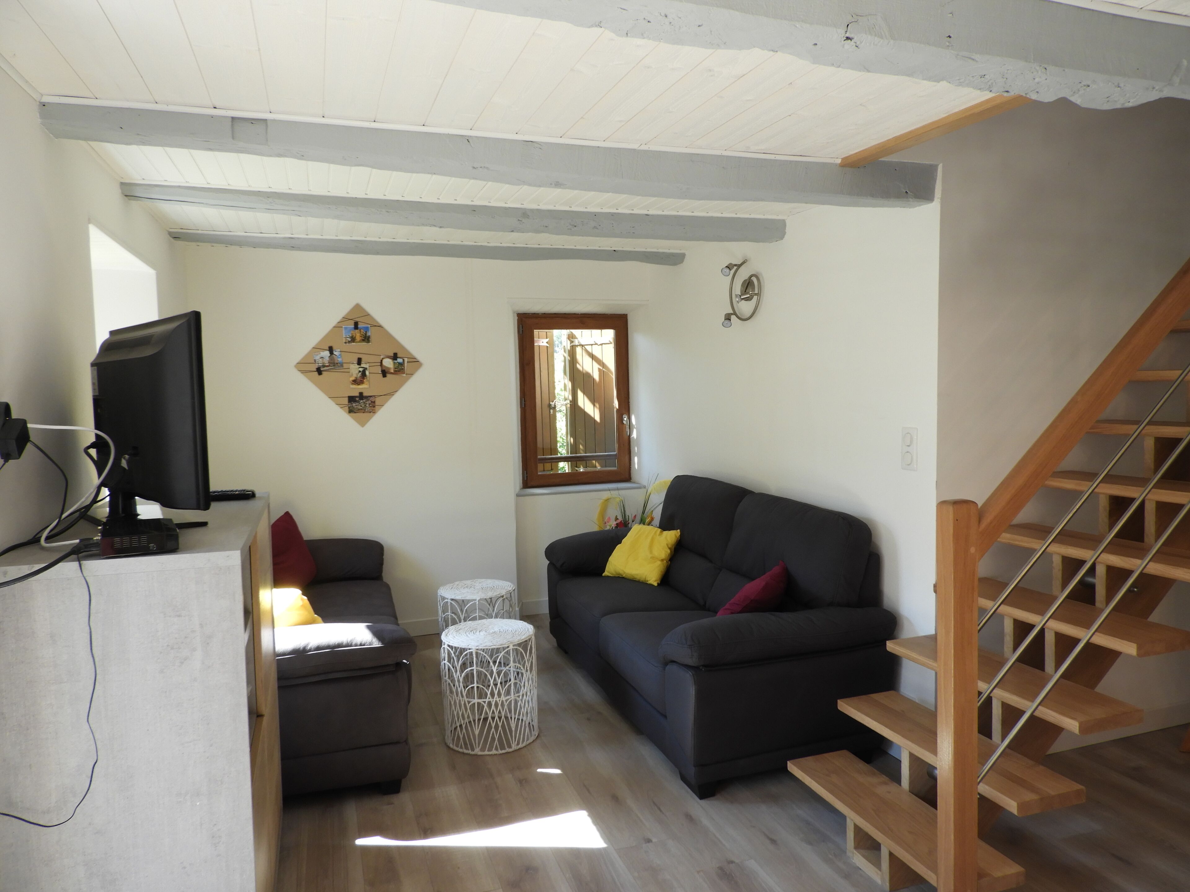 Una sala de estar con un sofá, un reposapiés y una televisión. Se ve una ventana con vistas a árboles y una escalera de madera.