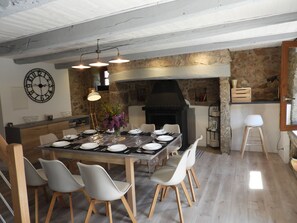 A dining area with a stone wall, a large clock, and a modern kitchen with a built-in grill. - Le Cabanel (Florentin-la-Capelle)
