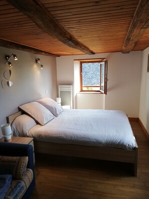 A bedroom with a wooden ceiling, a bed with white bedding, a bedside table with a lamp, and a window with a view. - Le Cabanel (Florentin-la-Capelle)