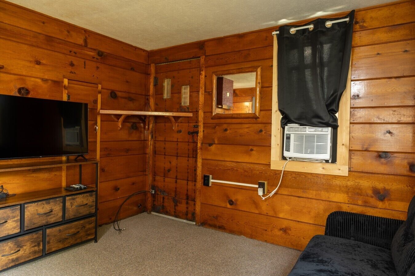 Une pièce aux murs en bois, un téléviseur pos é sur un meuble en bois, une fenêtre avec un appareil de climatisation et un rideau noir.