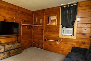 A room with wooden walls, a television on a wooden stand, a window with an air conditioning unit, and a black curtain.