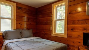 Une pièce de cabane en bois comprenant un lit, deux fenêtres et un téléviseur posé sur un meuble en bois.