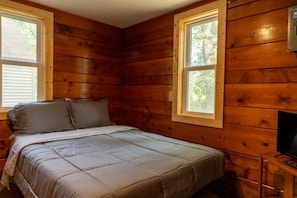 A wooden cabin room with a bed, two windows, and a television on a wooden stand.
