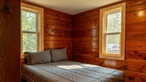Quarto de cabana de madeira com cama, duas janelas e vista para as árvores.