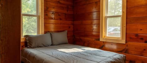 A wooden cabin room with a bed, two windows, and a view of trees.