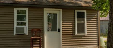 A single-story house with a front door, two windows, a wooden chair, and a small porch.