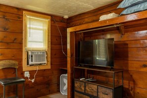 A wooden-paneled room with a television on a stand, a chair, a small table, and a window with blinds.