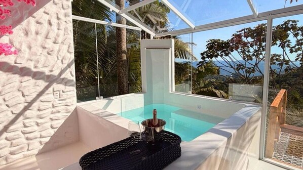 A glass-enclosed hot tub with a drink and glass, surrounded by a white wall and palm trees.