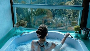 Una persona en una bañera con vista a la vegetación tropical a través de una ventana.
