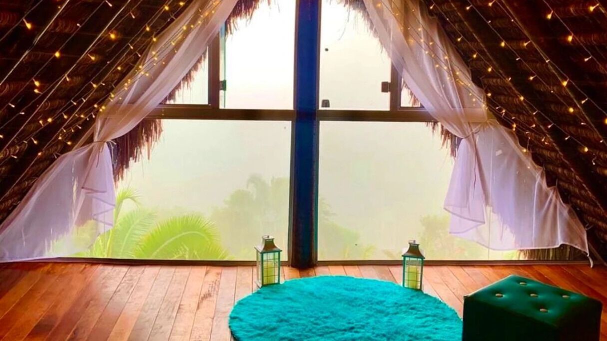 Una habitación con un ventanal grande, techo de paja y una alfombra redonda verde.