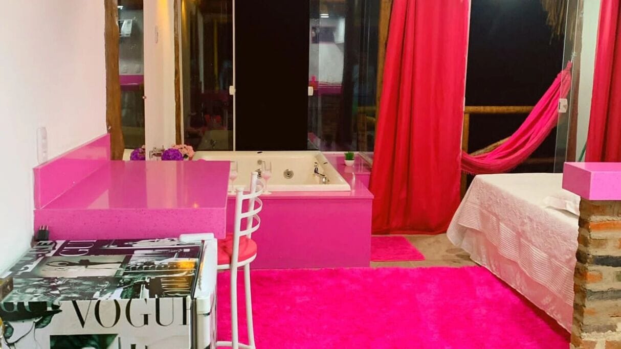 Une pièce avec une coiffeuse rose, une baignoire, un lit recouvert d’une couverture blanche et une fenêtre avec des rideaux rouges.