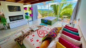 A modern bedroom with a large bed, a television, a bathtub, and a balcony with a view of the landscape.