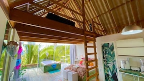 A room with a wooden ceiling, a bed, a small table, a chair, a window with a view of greenery, and a bathroom with a sink and a mirror.