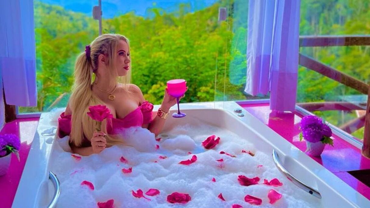 Una persona en una bañera llena de agua y pétalos de rosa, sosteniendo un vaso rosa.
