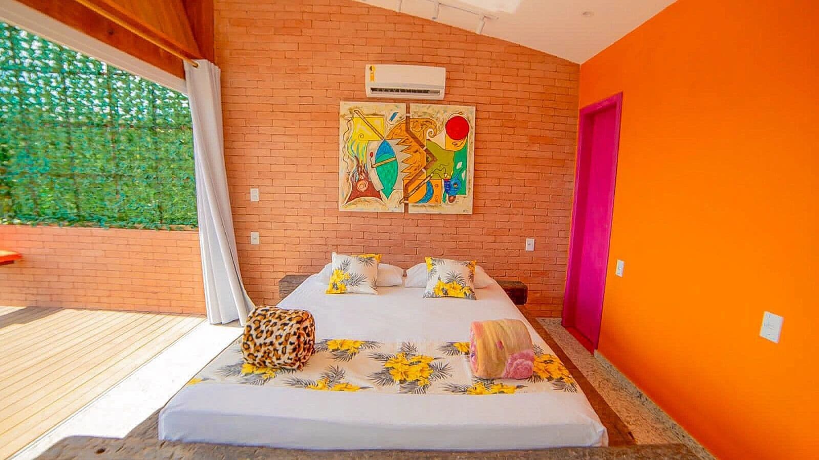 Quarto com cama, janela com cortinas, parede de tijolos e um quadro colorido.