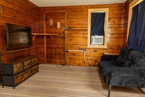 Ein Holzhäuschen-Interieur mit einem Fernseher auf einem Standfuß, einer Couch und einem Fenster mit Vorhängen.