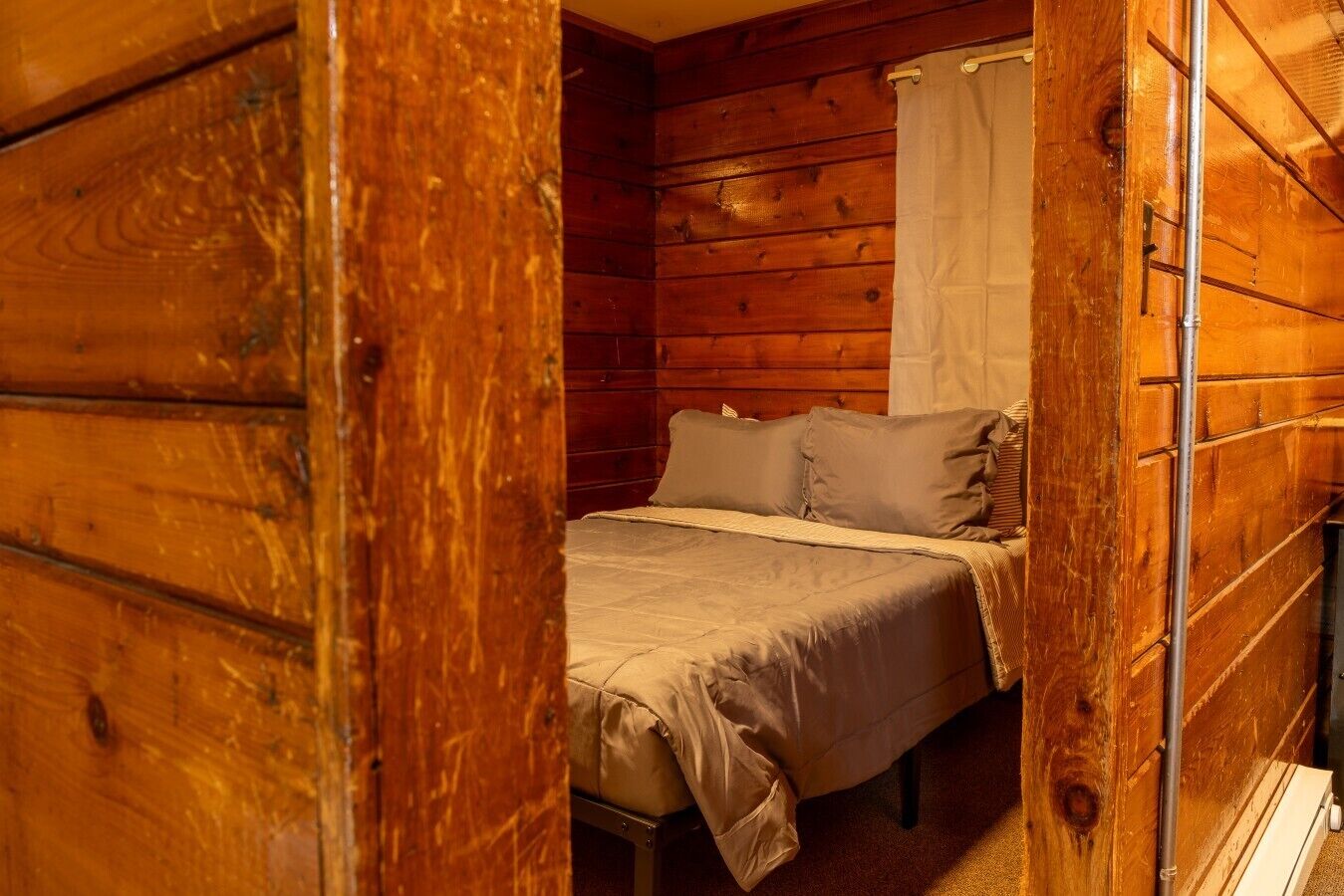 A wooden cabin interior with a bed, a window with a curtain, and a wooden wall.