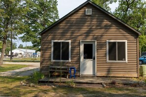Une petite cabane en bois d’un seul étage, avec une porte d’entrée et deux fenêtres, entourée d’arbres et bordée d’un chemin de gravier.