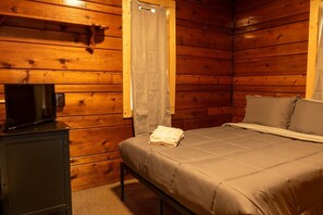 Une pièce de cabane en bois comprenant un lit, un téléviseur posé sur un meuble et une fenêtre avec un rideau.