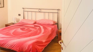 Una habitación con cama, suelo de madera, una mesita de noche y una lámpara fijada a la pared.