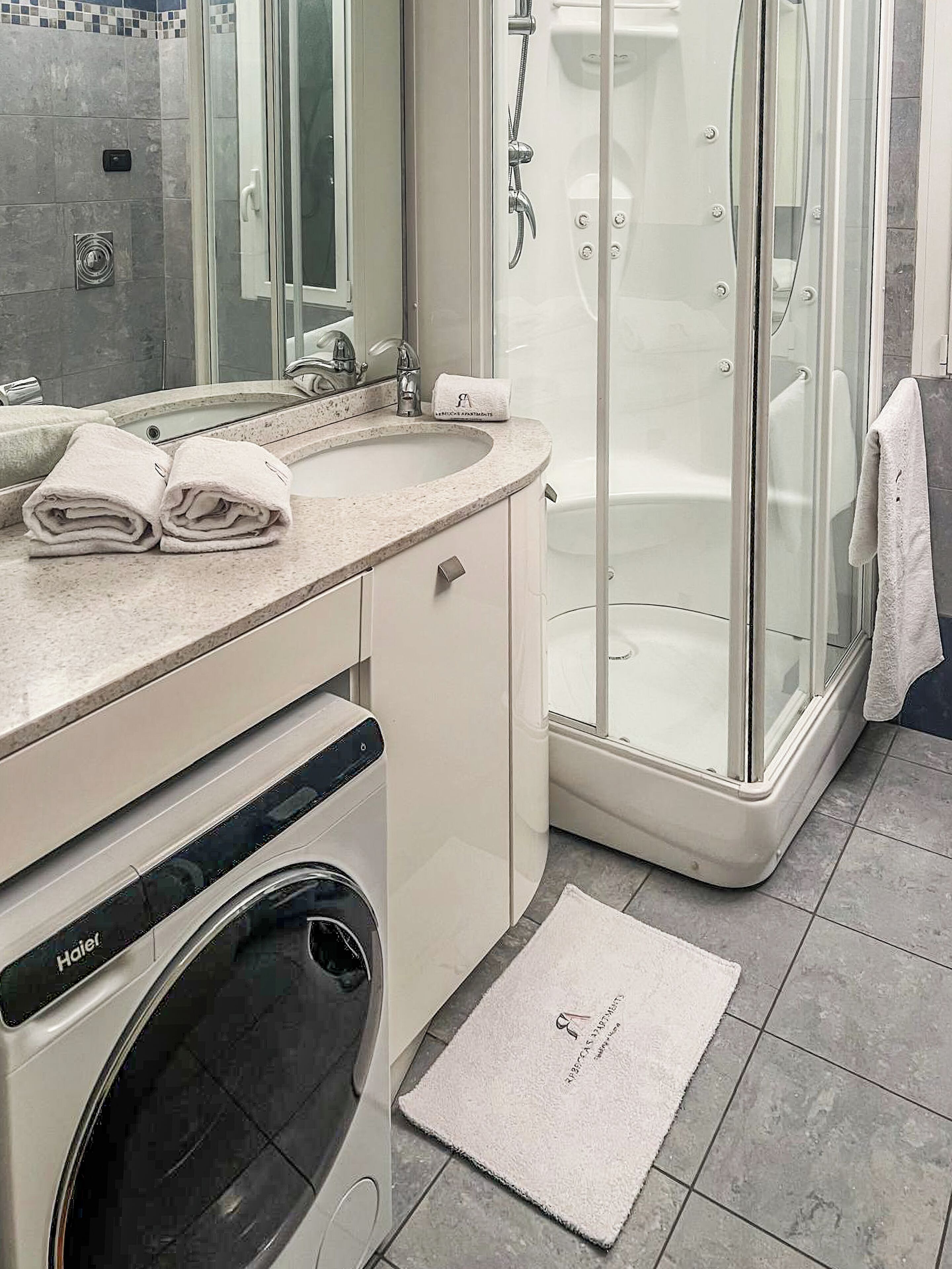 A modern bathroom with a built-in washing machine, a glass shower enclosure, and a vanity with a sink and towel rack.