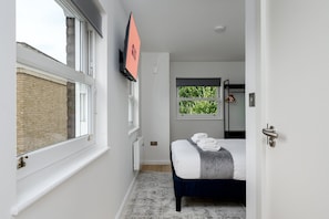 A narrow hallway leading to a bedroom with a bed, a radiator, and a window with a view of greenery. - GuestReady - Studio by the Maritime Museum (London)