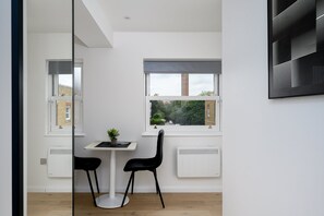 A room with a white table, two black chairs, a potted plant, and a window with a view of a tree and a building. - GuestReady - Studio by the Maritime Museum (London)